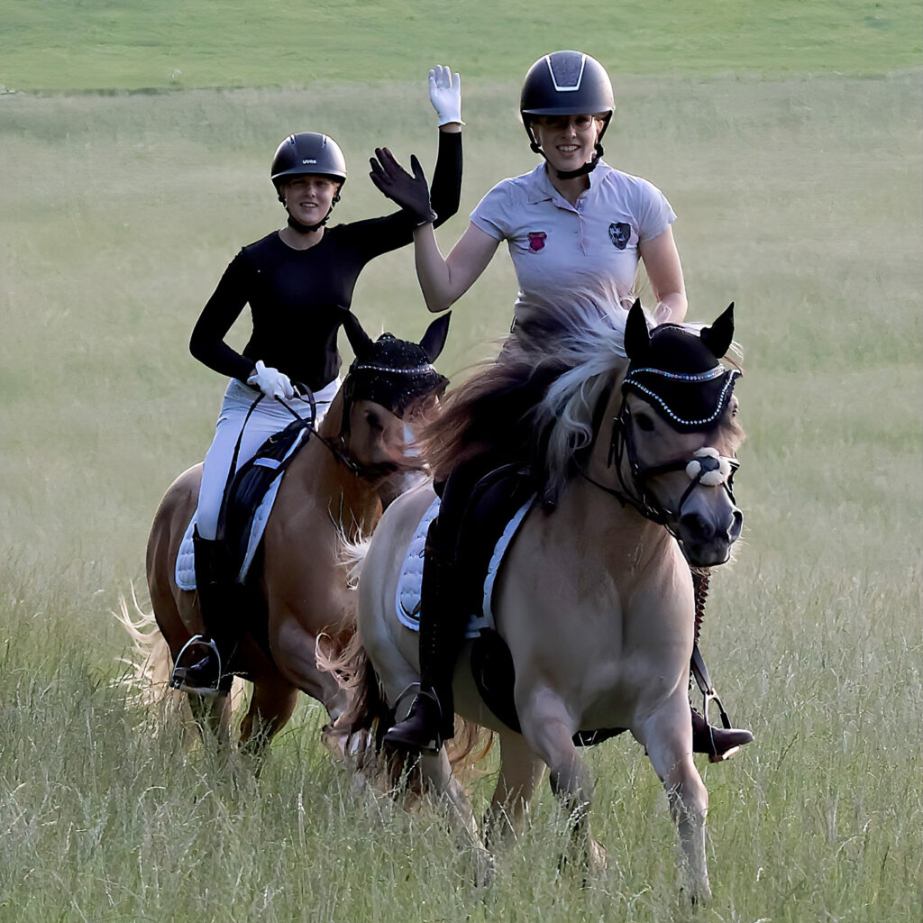 Zwei Reiterinnen reiten über eine Wiese und winken