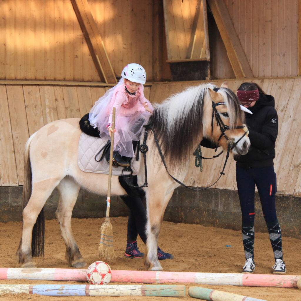 Ein als Prinzessin verkleidetes Kind versucht von einem Pony aus, einen Ball mit einem Besen zu verschieben